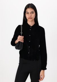 Mustast sametist koosneva nuppudega pikkade varrukatega särk, millel on terav krae ja läikivad nupud, koos väikese musta koti ja ahelkandjaga.