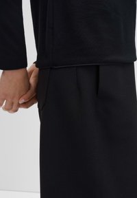 Schwarzes Langarmoberteil, kombiniert mit schwarzen Hosen. Das Oberteil hat eine gerippte Textur, während die Hosen eine glatte Oberfläche und Seitentaschen besitzen.
