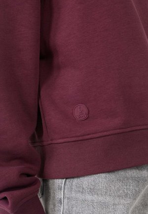 Bordeauxrode sweatshirt gemaakt van zacht materiaal, met een geribde tailleband en manchetten, en een subtiele logopatch in de benedenhoek.