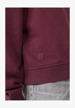 Sweatshirt bordeaux en tissu doux, avec une taille et des poignets côtelés, arborant un discret patch logo dans le coin inférieur.
