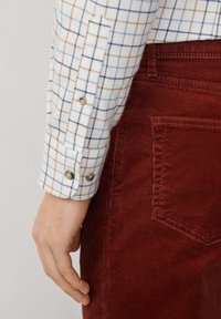 Camicia bianca a quadri con linee blu e marroni, con polsini bottonati; abbinata a pantaloni in corduroy color ruggine, che mettono in risalto la texture delle tasche.