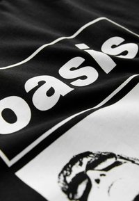 Tricou negru din bumbac cu text alb îndrăzneț "oasis" și un grafic al feței unei persoane în alb și negru, cu o textură a țesăturii ușor ridată.