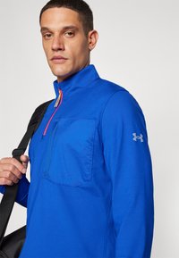 Blå zip-up pullover med høj krave, har en frontlomme, farverig lynlåsdetaljering og et logo på ærmet. Glat, letvægtsstof.
