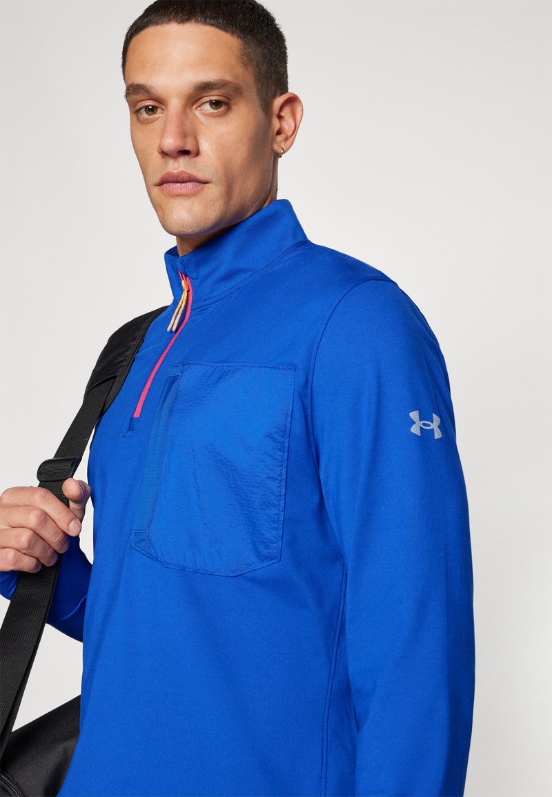 Sudadera azul con cremallera, de cuello alto, que cuenta con un bolsillo frontal, detalles de cremallera coloridos y un logo en la manga. Tela suave y ligera.