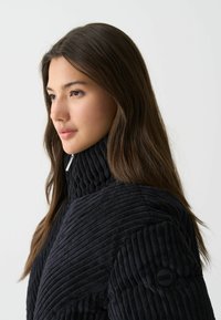 Zwarte textuurjacket met een hoge kraag en geribbelde stof. Heeft een ronde zwarte knop op de linkermouw. Glad haar is naar beneden gestyled.