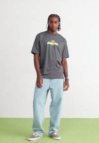 Grå bomulds t-shirt med et centralt logo design i lys grøn og rød, parret med lyseblå løstsiddende denim jeans og grå sneakers.