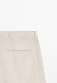 Pantalon beige à taille élastique avec une unique poche arrière fendue horizontale et des détails de couture minimaux.