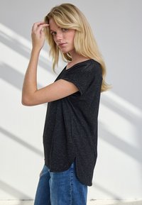 Blond naine mustas lühikeste varrukatega särgis ja sinistes teksapükstes puudutab juukseid, seistes valge seina ääres, millel on diagonaalsed varjud.