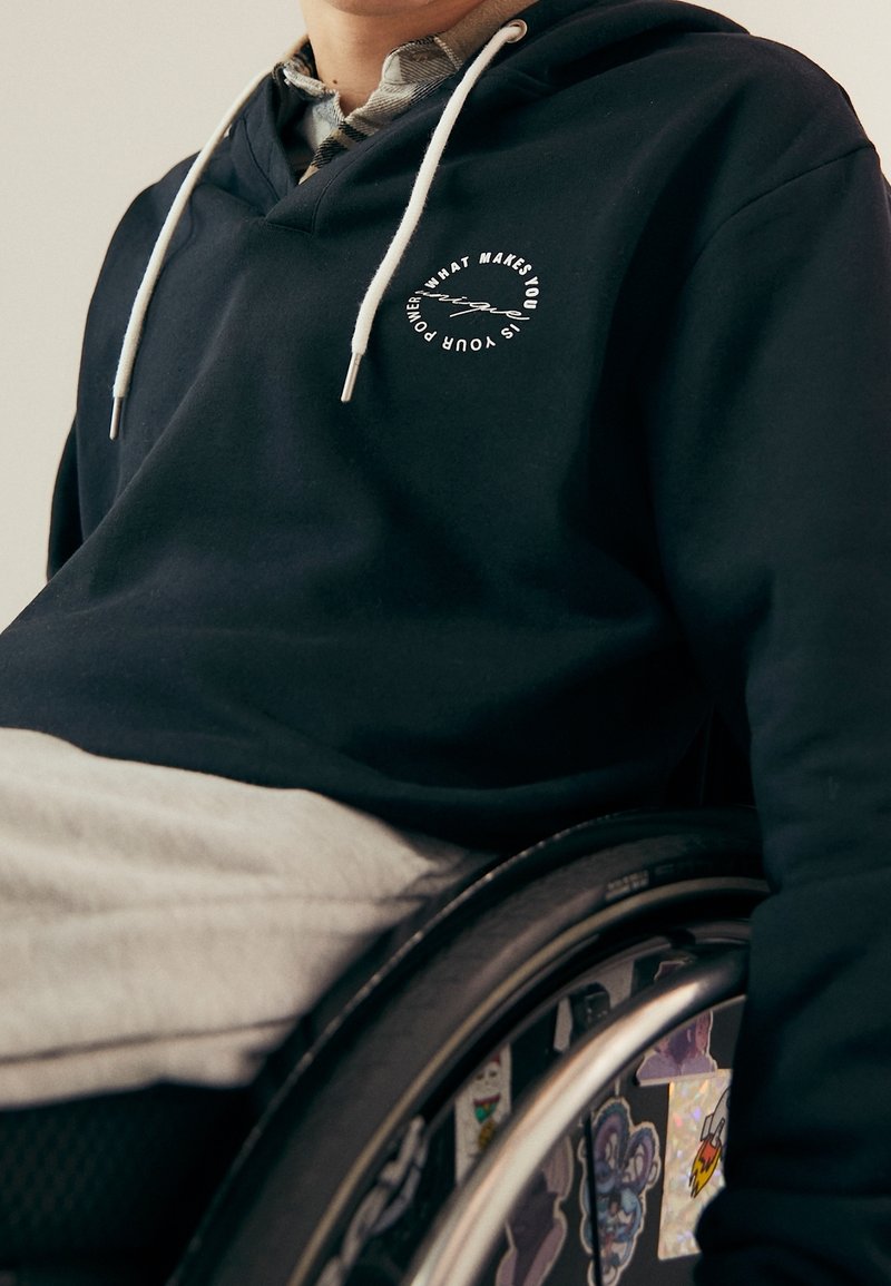 Felpa nera con logo testuale circolare bianco sul petto, coulisse, indossata sopra pantaloni della tuta grigi; persona su una sedia a rotelle.