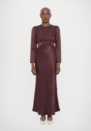 Robe en satin bourgogne longue avec des manches longues, des découpes au niveau de la taille et une silhouette lisse et fluide. Associée à des chaussures de couleur crème.