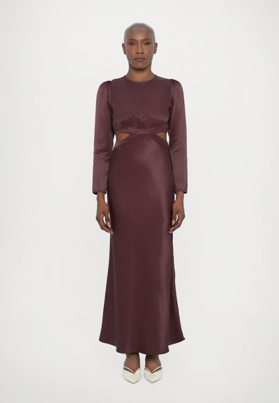 Robe en satin bourgogne longue avec des manches longues, des découpes au niveau de la taille et une silhouette lisse et fluide. Associée à des chaussures de couleur crème.