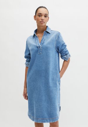 Kobieta z włosami zaczesanymi do tyłu, ubrana w jasnoniebieską sukienkę dżinsową z podwiniętymi rękawami, stojąca na jednolitym białym tle.