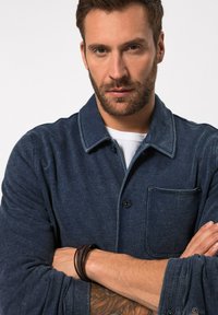 Námořnická modrá košile na knoflíky vyrobená z měkkého materiálu, s náprsní kapsou, zaobleným límcem a ohrnutými rukávy s viditelným švem.