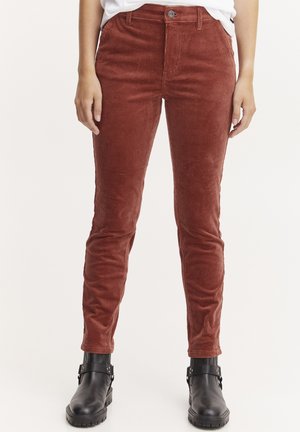 Pantalons en velours côtelé orange brûlé avec une coupe slim, présentant des côtes verticales, une fermeture à bouton et des poches, associés à des bottines noires.