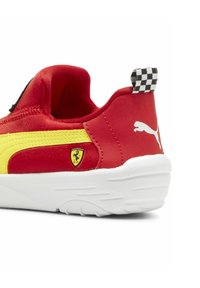 Punainen lenkkari, jossa on keltainen raita, teksturoitu kangas, valkoinen pohja, mustat yksityiskohdat ja ruudullinen vetonauha. Varustettu pienellä Ferrari-logolla.