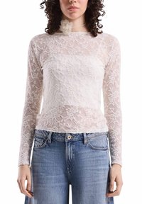 Weißes Spitzentop mit langen Ärmeln, floraler Stickerei und semi-transparentem Material, kombiniert mit hellblauen Jeans.
