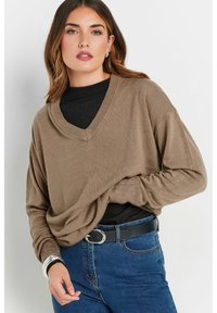 Brauner, übergroßer V-Ausschnitt-Pullover mit langen Ärmeln, aus einem weichen Stoff. Stylisch über einem schwarzen Oberteil getragen und kombiniert mit hoch taillierten blauen Jeans.