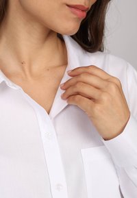Chemise blanche à boutons, fabriquée en tissu lisse, dotée d'un col classique, d'une poche avant et de manches longues avec poignets boutonnés.
