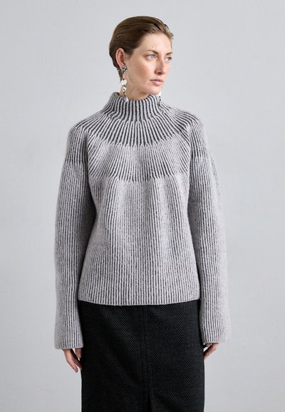 Světle šedý pletený svetr s ribbed texturou, vysokým límcem a širokými rukávy. Kombinován s černou texturovanou sukní.