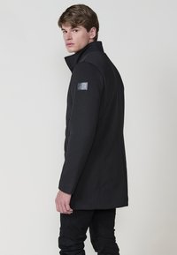 Abrigo negro con un alto cuello, cierre de un solo botón y una textura sutil. Presenta un parche de logo en el costado y se combina con pantalones negros.