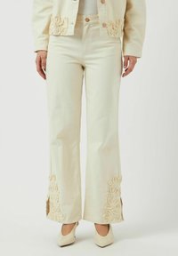 Pantalones de mezclilla color crema acampanados con bordados florales decorativos en color crema en el dobladillo y aberturas laterales. Botones en la cintura.