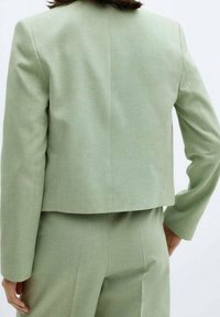 Blazer court vert clair en tissu texturé, avec des manches longues et une silhouette épurée et ajustée. Vue de dos montrant des détails de design minimalistes.
