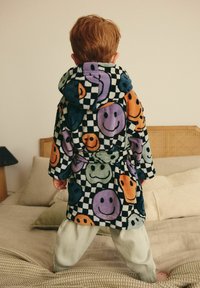 Fleece-Bademantel mit einem schwarz-weißen karrierten Muster, das farbenfrohe Smiley-Gesichter zeigt. Kapuzenstil mit einem gebundenen Gürtel, kombiniert mit hellen Hosen.