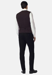 Brązowa kamizelka z tkaniny z zapięciem z tyłu, noszona na białej koszuli, w połączeniu z czarnymi spodniami, widok z tyłu.