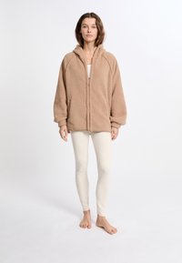 Giacca in fleece beige con cappuccio, dotata di zip frontale, maniche raglan e una texture morbida, indossata sopra leggings chiari.