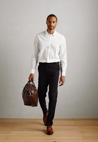 Hombre vestido con una camisa blanca, pantalones negros y zapatos marrones, caminando sobre un suelo de madera, llevando un gran bolso de cuero marrón en la mano derecha.