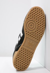 Zwarte skate schoen met witte strepen, bovenwerk van textuur-suède en rubberen gumzool met een honingraatpatroon en Adidas-logo.