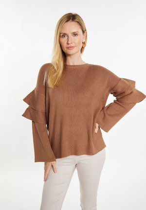 Brauner gestrickter Pullover mit Rüschensleeves und lockerer Passform. Strukturierter Stoff und Rundhalsausschnitt. Kombiniert mit hellbeigen Hosen.