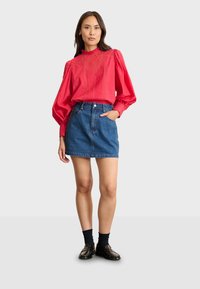 Blusa roja de manga larga con cuello rizado y detalle texturizado, combinada con una falda corta de mezclilla azul y mocasines negros con calcetas.