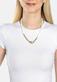 Vitt pärlhalsband med guldmetallackenter, med ett ovalt lås och växelvis släta och strukturerade mönster, bärs över en vit skjorta.