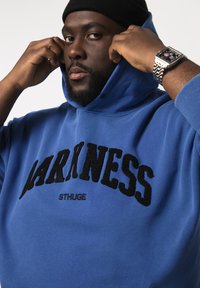 Niebieska bluza z kapturem, wykonana z miękkiego materiału, z odważnym czarnym napisem "DARKNESS" oraz logo "STHUGE". Kapucha jest częściowo naciągnięta, a szwy są widoczne.