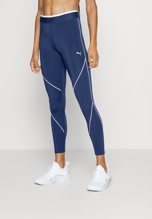 Leggings de sport bleu marine avec des détails en piping blanc, taille haute et design ajusté. Portés avec des chaussures de sport blanches.