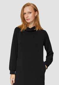 Zwarte jurk met lange mouwen en een cowl halslijn, gemaakt van een gladde stof. Heeft zijzakken en taps toelopende manchetten. Eenvoudige, rechte silhouet.