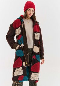 Žena oblečená v farebnom patchwork fleecovom kabáte, červené pletenej čiapke, béžovom svetri a nohaviciach s leopardím vzorom stojí na jednolítom pozadí.