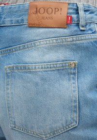 Svetlo modri žep na zadnji strani jeans hlač z rjavim usnjenim etiketom blagovne znamke Joop! Jeans, všitim nad žepom.