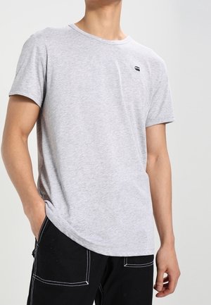 Osoba ubrana w jasnoszary t-shirt z krótkim rękawem oraz czarne spodnie z białym przeszyciem, jedną ręką w kieszeni, na jednolitym tle.