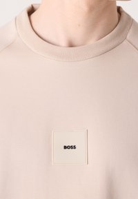 Sweatshirt beige avec un col ras du cou côtelé. Présente un écusson carré avec le logo dans une teinte plus claire et le mot "BOSS" en noir. Texture lisse.