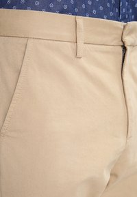 Beige trousers with a smooth, durable fabric. Features include front pockets, a zip fly, and a belt loop at the waistband.