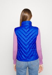 Blue, chevron-patterned puffer vest with high collar; worn over a pink long-sleeve top, paired with blue denim jeans.