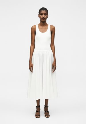 Kvinne iført en hvit ermeløs ribbestrikket topp og en flytende hvit midiskjørt, kombinert med svarte høye hæler med stropper, stående rett.