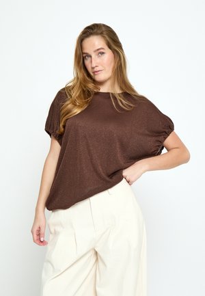 Kobieta o długich blond włosach, ubrana w luźną brązową bluzkę z krótkim rękawem i kremowe, szerokie spodnie z wysokim stanem, pozuje z ręką na biodrze.