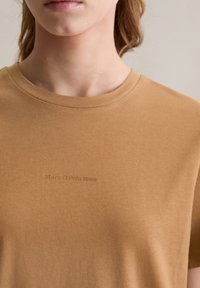 Braunes Baumwoll-T-Shirt mit Rundhalsausschnitt, das ein dezentes Logo "Marc O'Polo DENIM" in einer dunkleren Farbe aufgedruckt hat. Glatte Textur.