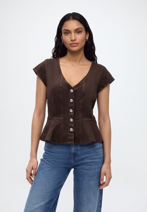Femme aux cheveux bruns ondulés porte un top marron cintré à boutons avec des manches courtes et un jean bleu sur un fond blanc uni.