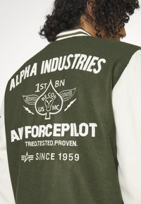 Giacca varsity verde con maniche bianche. Presenta testo ricamato, un logo a teschio e colletto a righe. Tessuto resistente con una vestibilità strutturata.
