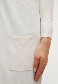 Ljusgrå stickad tröja med en framficka, ribbade muddar och en mjuk textur. Designen är avslappnad och funktionell.