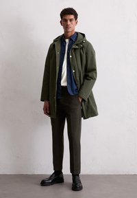 Mężczyzna w oliwkowej, zapinanej na zamek kurtce z kapturem, nałożonej na niebieską koszulę dżinsową i białą bluzkę, w ciemnych spodniach i czarnych mokasynach, stojący przy gładkiej ścianie.